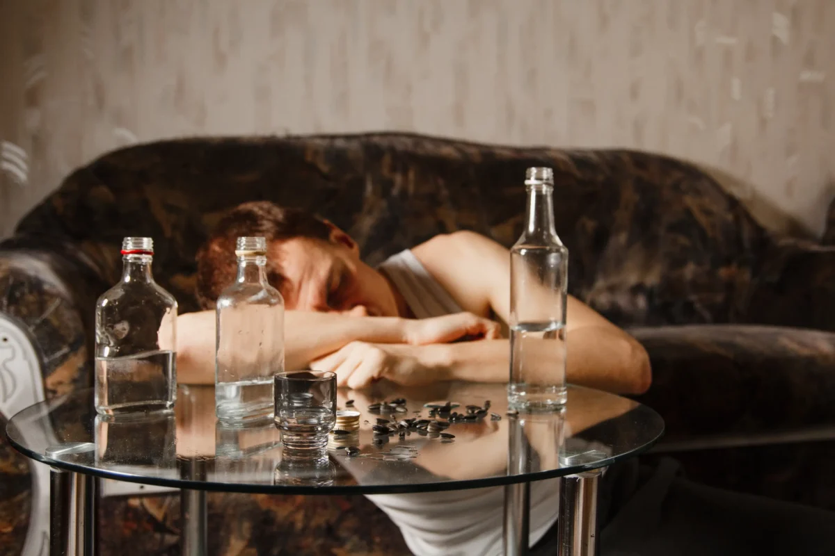 shutterstock_1373744261-změněno-na-3000x2000 | TVGURU.cz Osoba si opírá hlavu o paži přes skleněný stůl s lahvemi a mincemi rozházenými poblíž