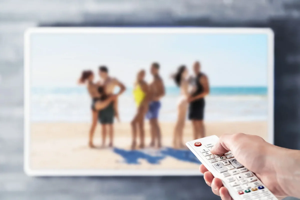 ruka s ovladačem namířená na televizi, kde je parta lidí na pláži
