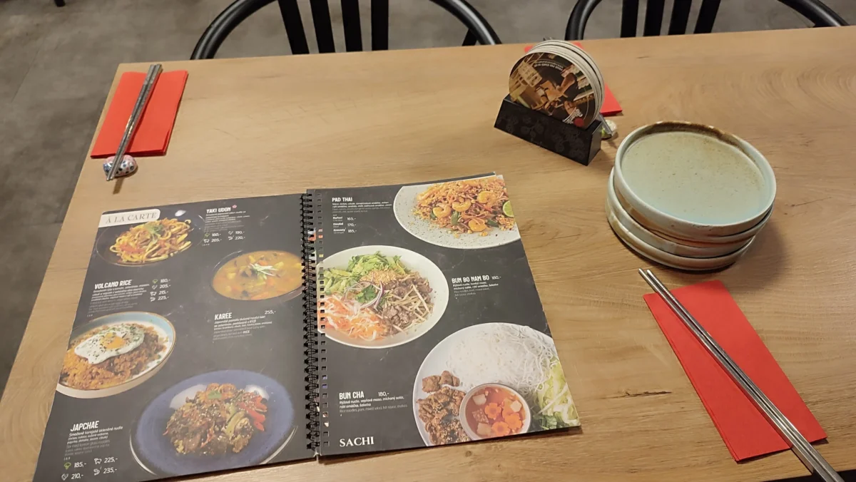 shutterstock_2691039709 1 | TVGURU.cz Open two-page menu on a wooden table with photos of dishes, chopsticks, and red napkins nearby.