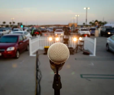 Close-up of a microphone on a stand in the foreground with a blurred outdoor parking lot full of cars in the background.