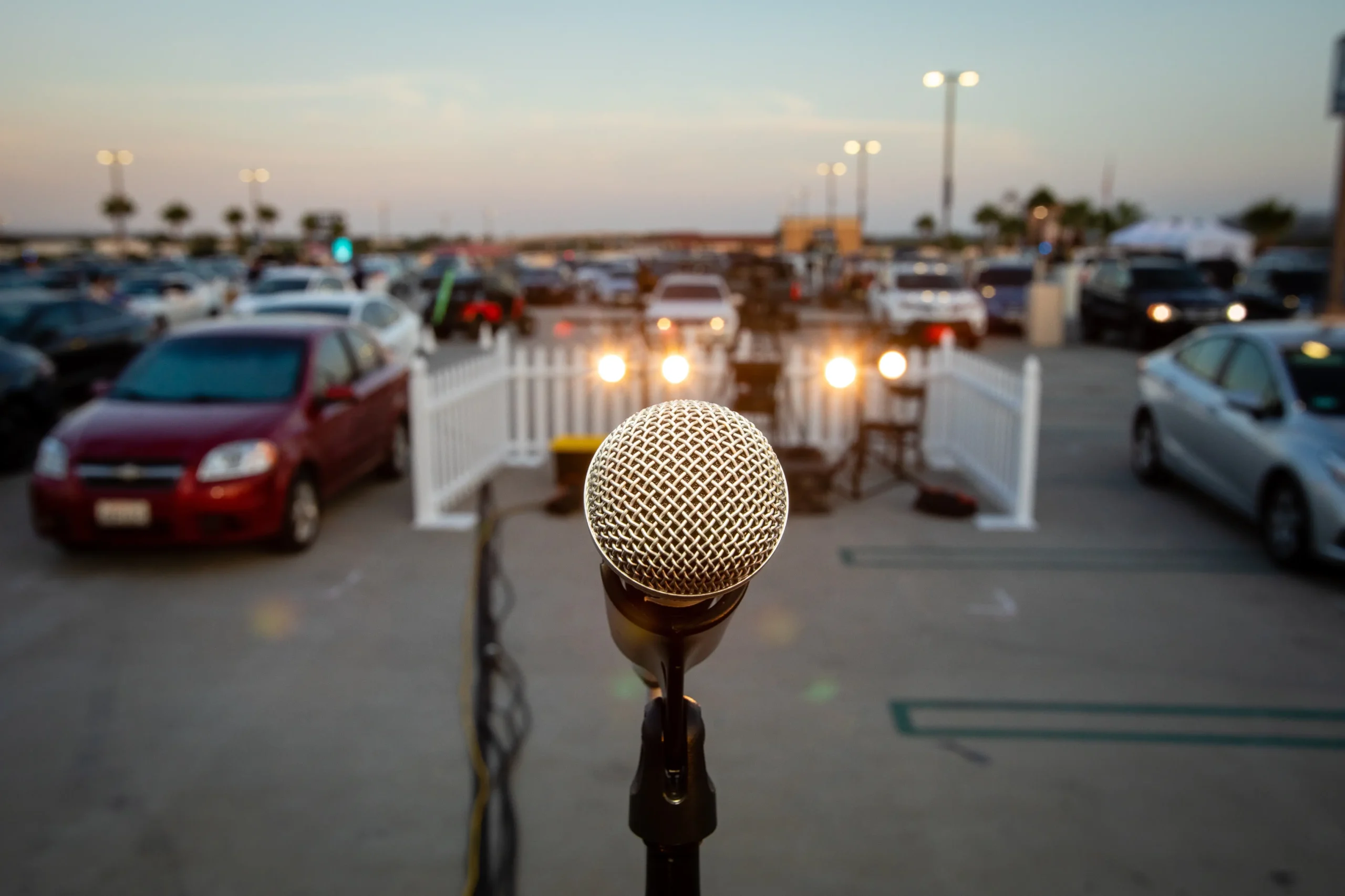 Close-up of a microphone on a stand in the foreground with a blurred outdoor parking lot full of cars in the background.