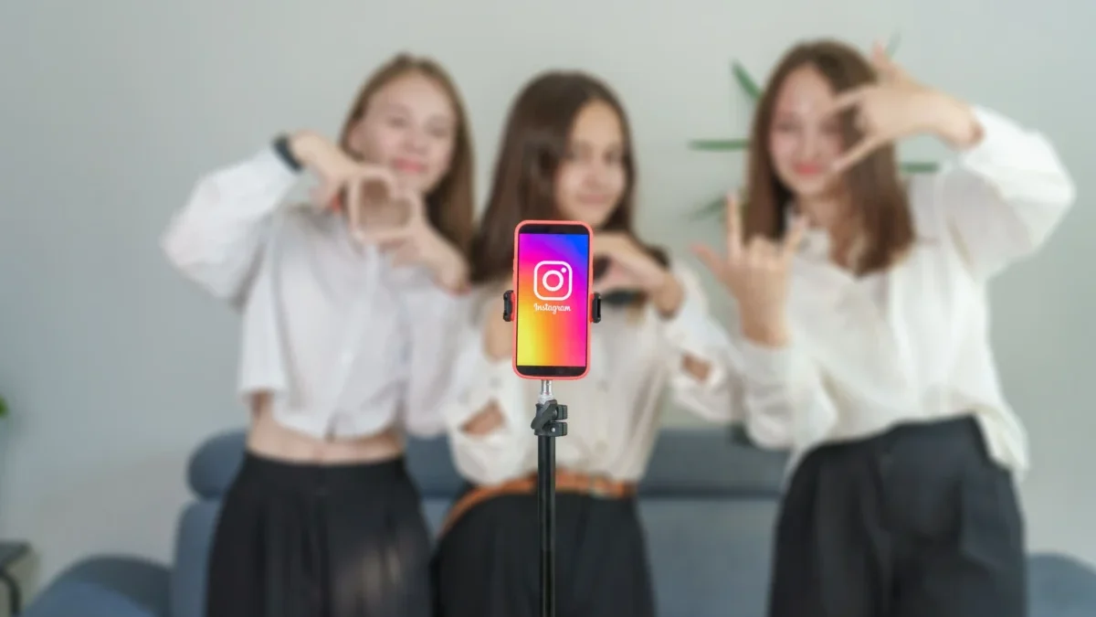 Four young women pose behind a smartphone on a selfie stick, centered on the Instagram app icon. Subtle, blurred background.