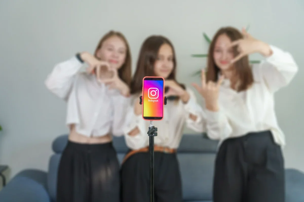 Four young women pose behind a smartphone on a selfie stick, centered on the Instagram app icon. Subtle, blurred background.