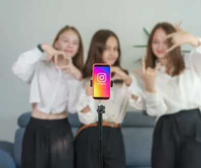 Four young women pose behind a smartphone on a selfie stick, centered on the Instagram app icon. Subtle, blurred background.