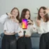 Four young women pose behind a smartphone on a selfie stick, centered on the Instagram app icon. Subtle, blurred background.