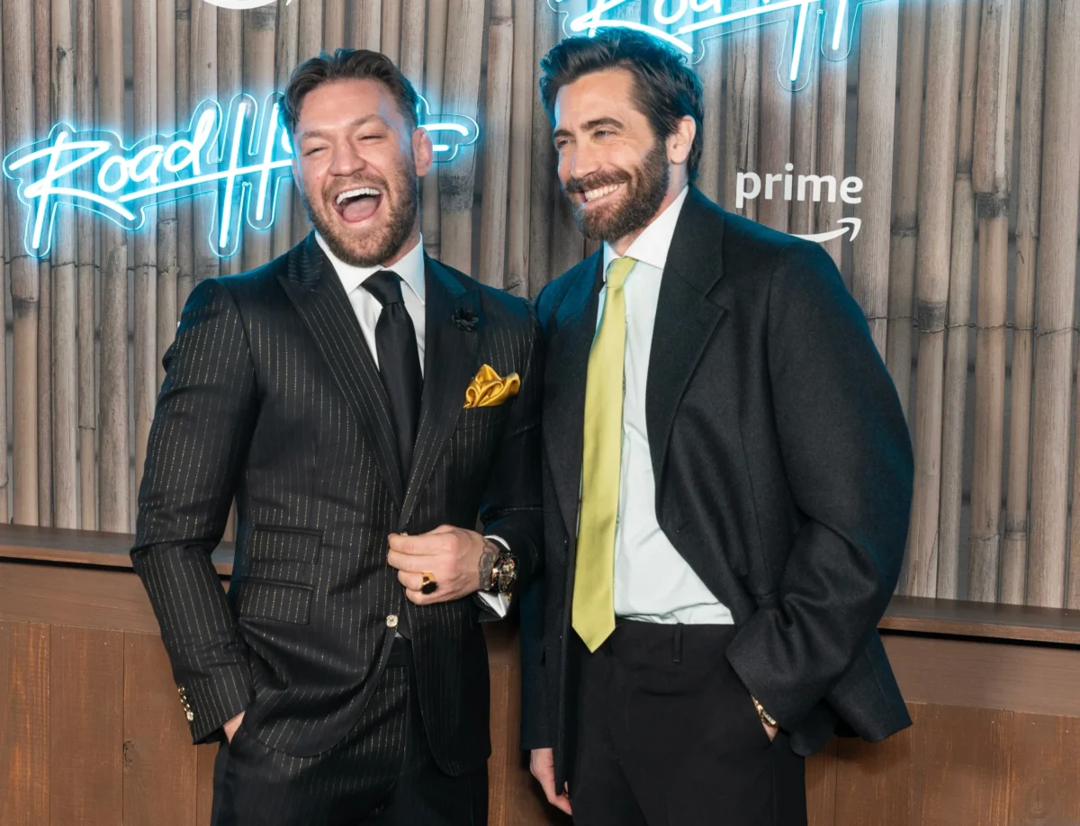 Dva muži v tmavých oblecích pózují a smějí se u dřevěné zdi s modrými neonovými nápisy za nimi. Jake Gyllenhaal a Conor McGregor