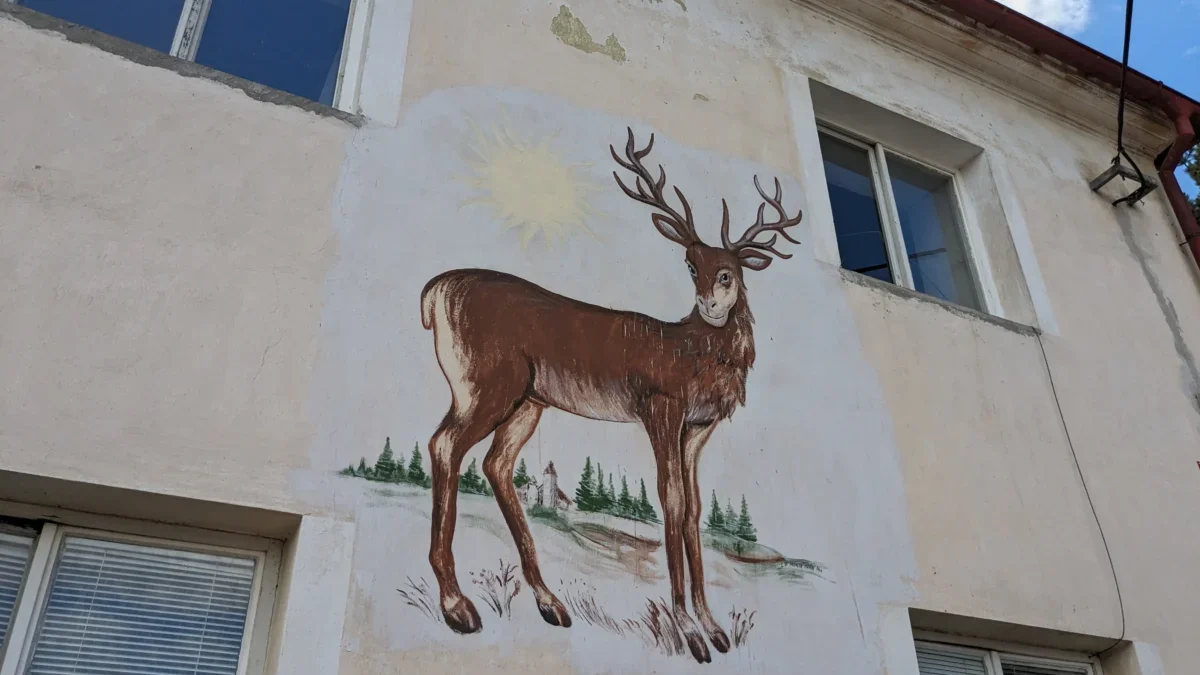 Exterior wall mural of a brown deer with large antlers, sun in the background, and pine trees painted beneath; building windows visible nearby.