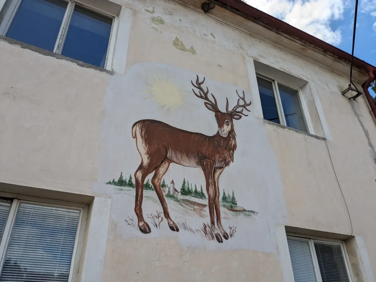 Exterior wall mural of a brown deer with large antlers, sun in the background, and pine trees painted beneath; building windows visible nearby.