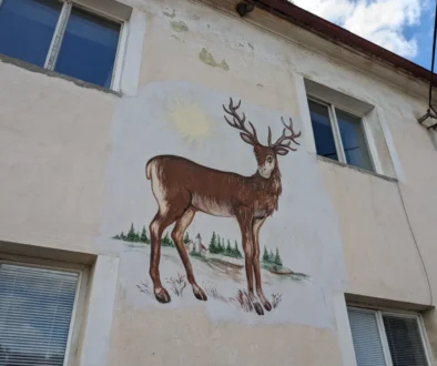 Exterior wall mural of a brown deer with large antlers, sun in the background, and pine trees painted beneath; building windows visible nearby.