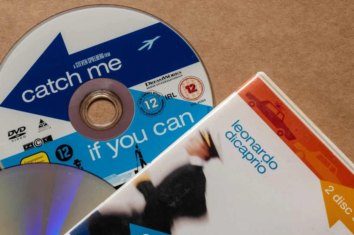 Detail filmového DVD s modrým obalem a nápisem „chyť mě, když to dokážeš“ a dalšími disky částečně viditelnými poblíž v nakloněném rozložení