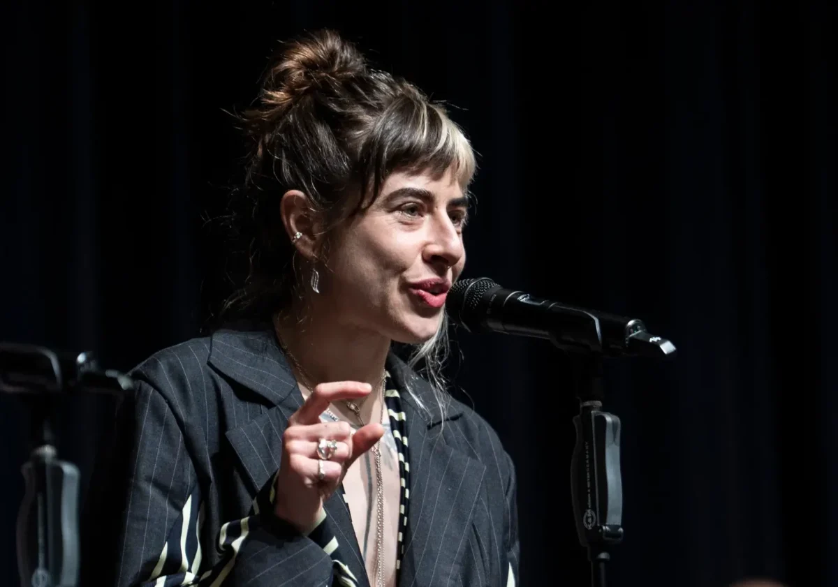 Female singer performing on stage, speaking into a microphone with a dark backdrop behind her.