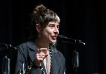 Female singer performing on stage, speaking into a microphone with a dark backdrop behind her.