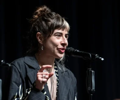 Female singer performing on stage, speaking into a microphone with a dark backdrop behind her.