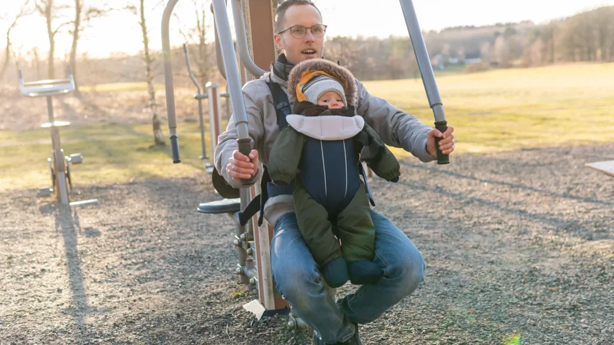 Father wearing glasses helps a toddler secured in a front-facing baby carrier on an outdoor playground structure at sunset/morning light.