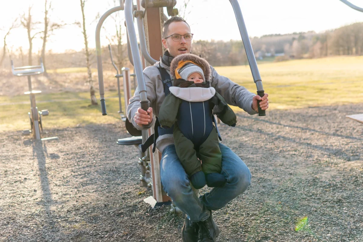 Father wearing glasses helps a toddler secured in a front-facing baby carrier on an outdoor playground structure at sunset/morning light.
