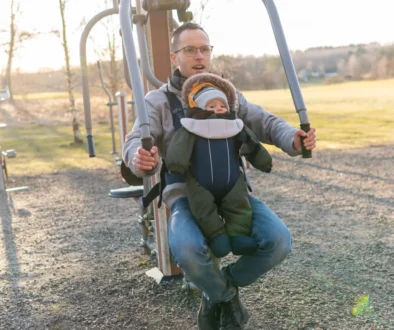 Father wearing glasses helps a toddler secured in a front-facing baby carrier on an outdoor playground structure at sunset/morning light.