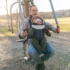 Father wearing glasses helps a toddler secured in a front-facing baby carrier on an outdoor playground structure at sunset/morning light.