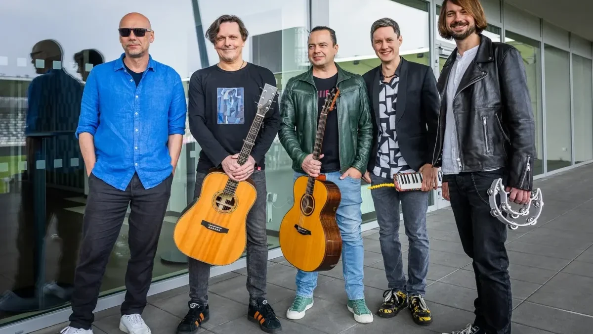 Five male musicians pose outside a glass-fronted building; two hold acoustic guitars, one has a small keyboard, and another has a tambourine in hand.