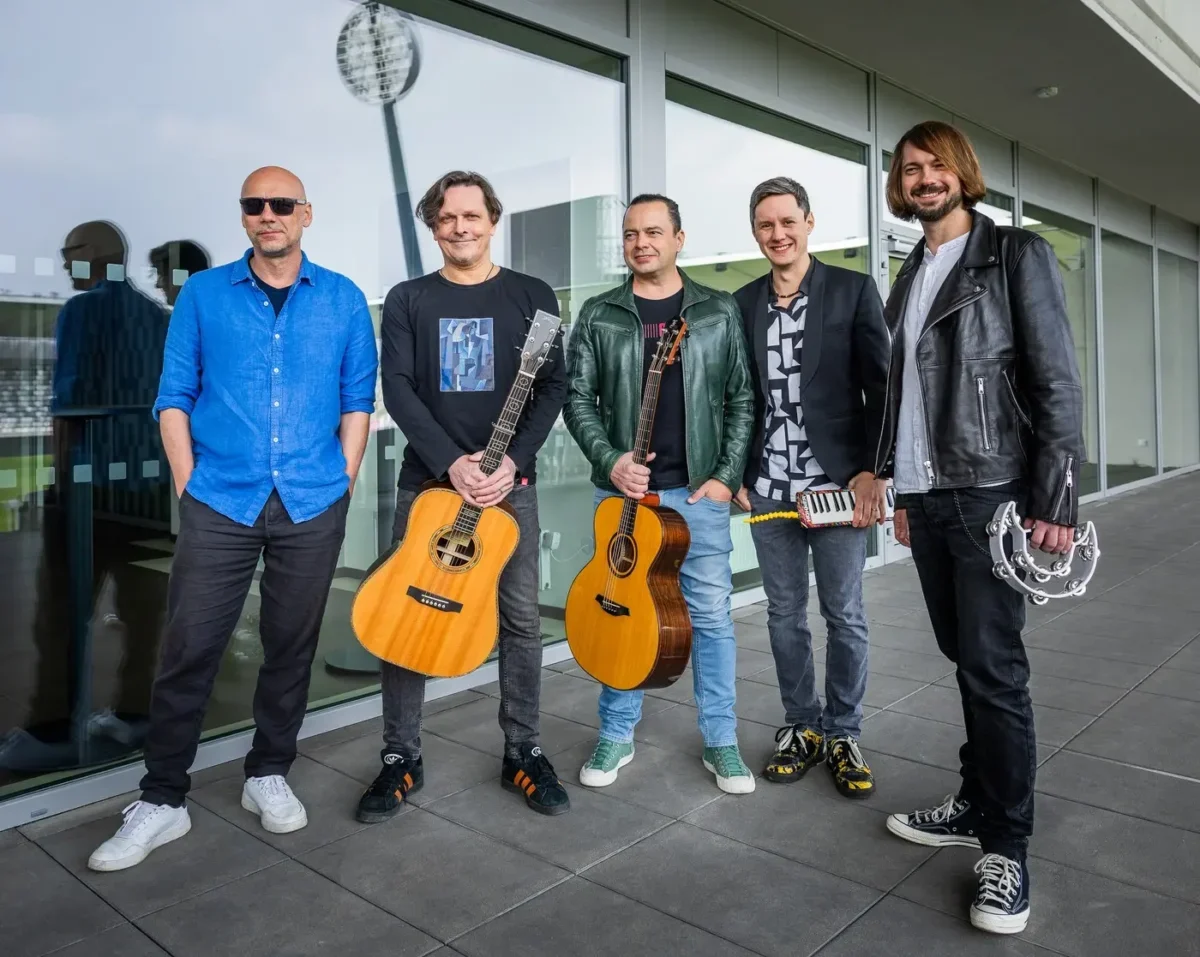 Five male musicians pose outside a glass-fronted building; two hold acoustic guitars, one has a small keyboard, and another has a tambourine in hand.
