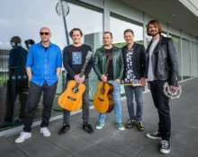 Five male musicians pose outside a glass-fronted building; two hold acoustic guitars, one has a small keyboard, and another has a tambourine in hand.