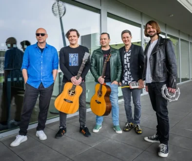 Five male musicians pose outside a glass-fronted building; two hold acoustic guitars, one has a small keyboard, and another has a tambourine in hand.