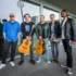 Five male musicians pose outside a glass-fronted building; two hold acoustic guitars, one has a small keyboard, and another has a tambourine in hand.
