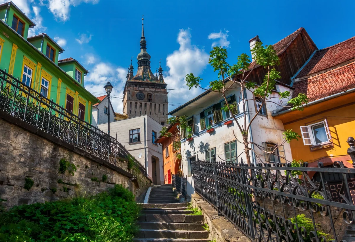 shutterstock_2759577171 1 | TVGURU.cz Stone steps and a black wrought-iron railing wind uphill between bright green-and-yellow houses toward a tall ornate clock tower under a blue sky.