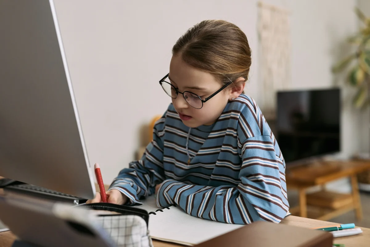 shutterstock_2760492267 1 | TVGURU.cz Child wearing glasses and a striped blue shirt draws with a red pencil at a desk beside a laptop and notebook.