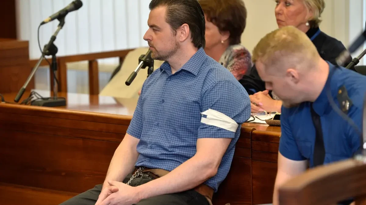 Muž v krátkém tričku s límečkem a s krátkými černými vlasy sedí u soudu a vedle něj sedí policista