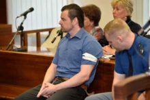 Muž v krátkém tričku s límečkem a s krátkými černými vlasy sedí u soudu a vedle něj sedí policista