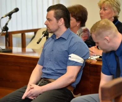 Muž v krátkém tričku s límečkem a s krátkými černými vlasy sedí u soudu a vedle něj sedí policista