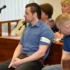 Muž v krátkém tričku s límečkem a s krátkými černými vlasy sedí u soudu a vedle něj sedí policista