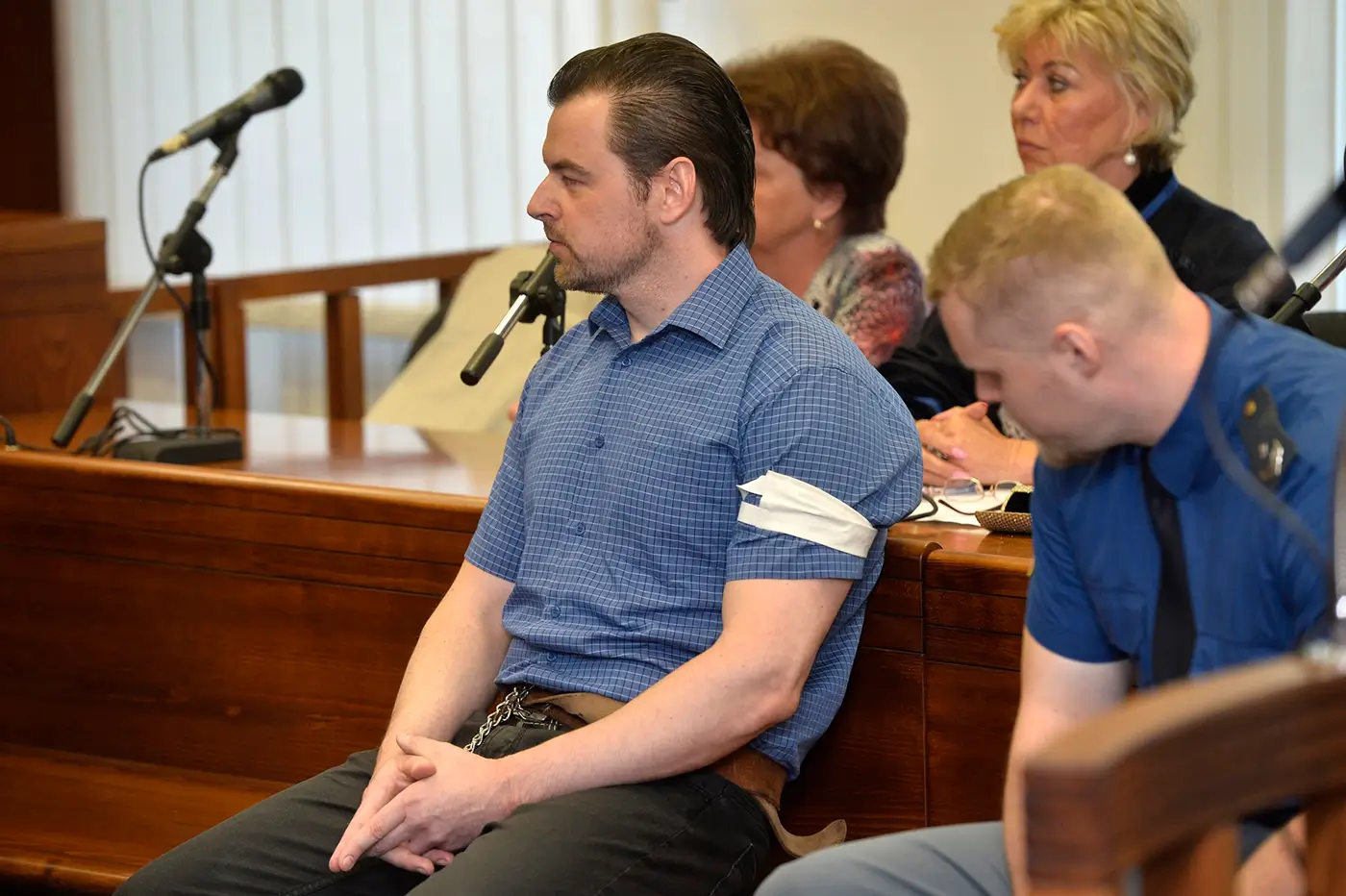 Muž v krátkém tričku s límečkem a s krátkými černými vlasy sedí u soudu a vedle něj sedí policista