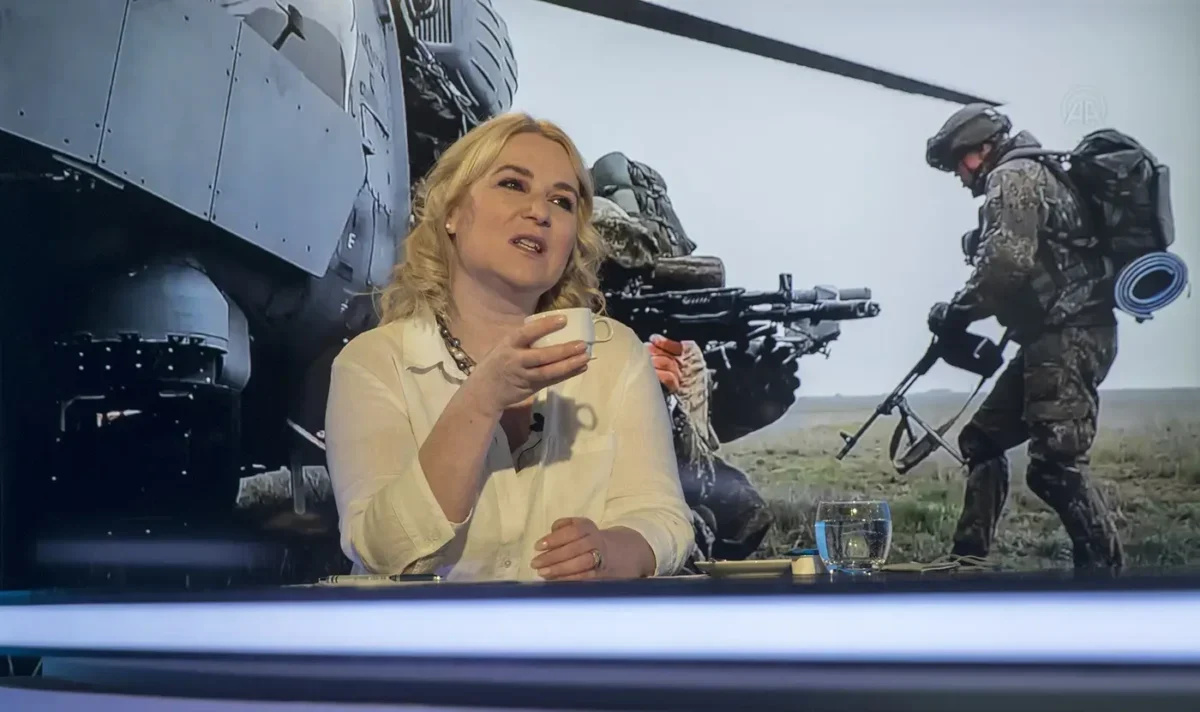 Žena se světlými vlasy sedí ve studiu a za ní se promítají vojenské záběry