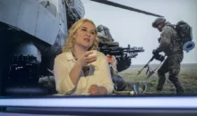 Žena se světlými vlasy sedí ve studiu a za ní se promítají vojenské záběry