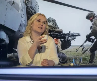 Žena se světlými vlasy sedí ve studiu a za ní se promítají vojenské záběry
