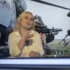 Žena se světlými vlasy sedí ve studiu a za ní se promítají vojenské záběry