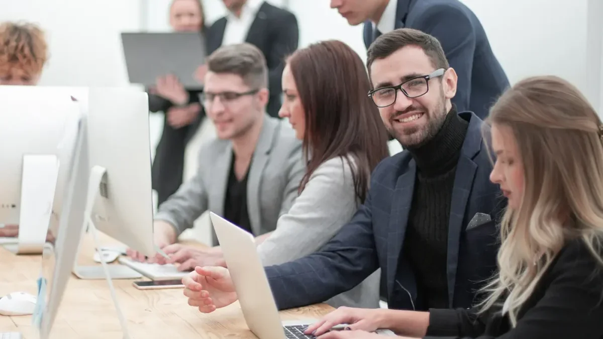 Skupina zaměstnanců, žen a mužů, ve společenském oblečení se shromažďují u pracovního stolu