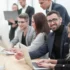 Skupina zaměstnanců, žen a mužů, ve společenském oblečení se shromažďují u pracovního stolu