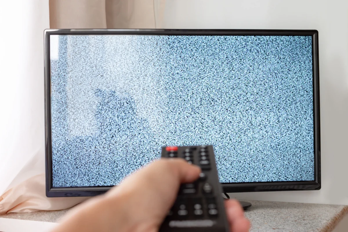 shutterstock_1303315147 1 | TVGURU.cz Person holding a TV remote and aiming it at a television displaying static/no signal.