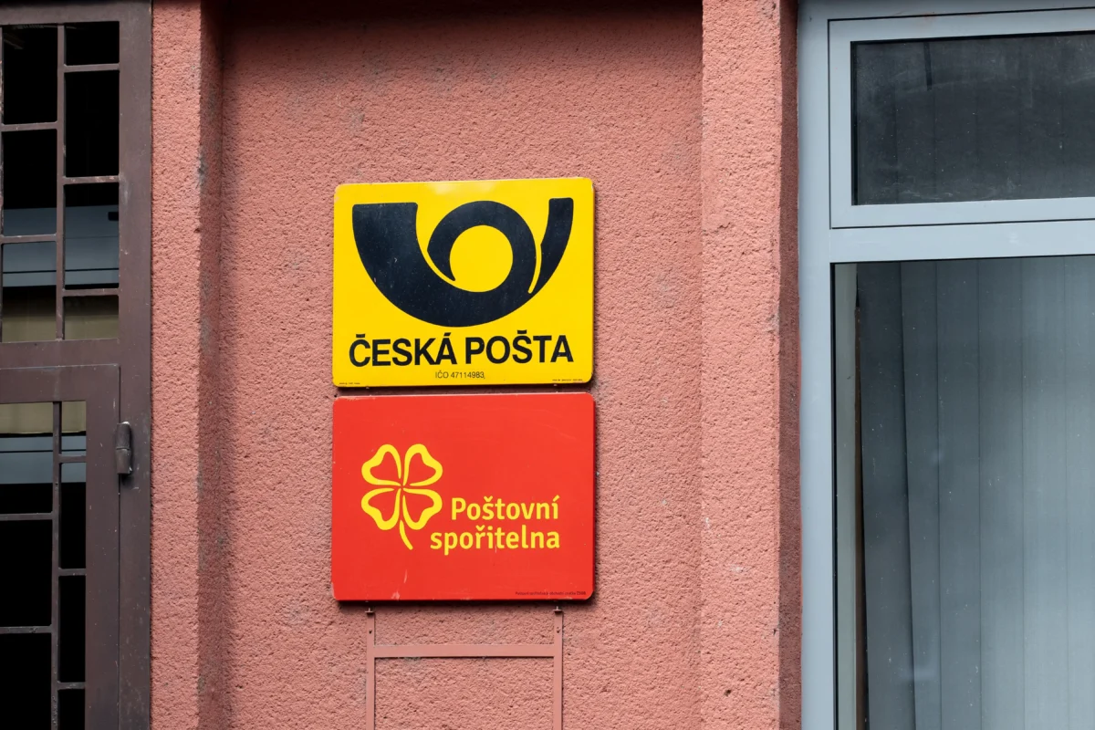 Yellow Česká Pošta sign with black logo mounted on a pink wall next to a window and metal grille