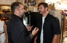 Dva muži v tmavém oblečení spolu hovoří v nějakém podniku, kde prodávají víno a pečivo