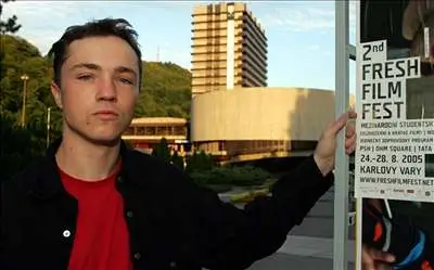 GRAd53c4_nemecek 1 | TVGURU.cz Young man in a red shirt and black jacket poses outdoors next to a poster for the 2nd Fresh Film Fest.