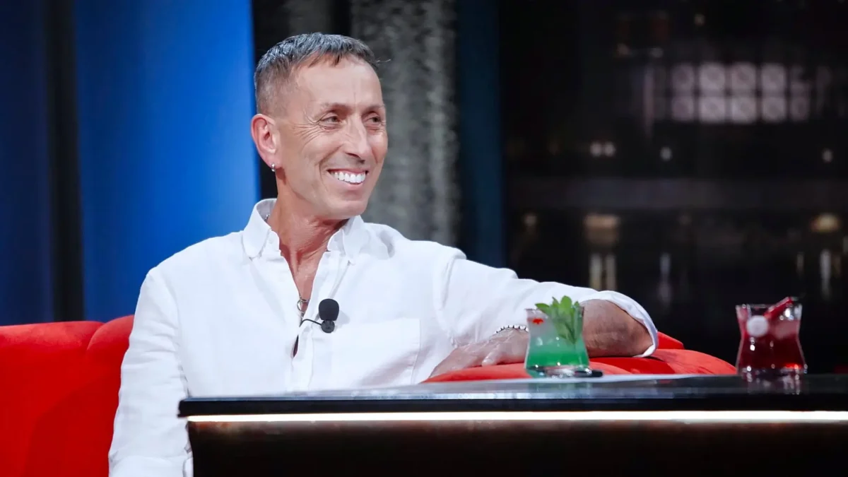 Smiling man in a white shirt on a talk-show set, sitting on a red couch with a microphone clipped to his shirt.