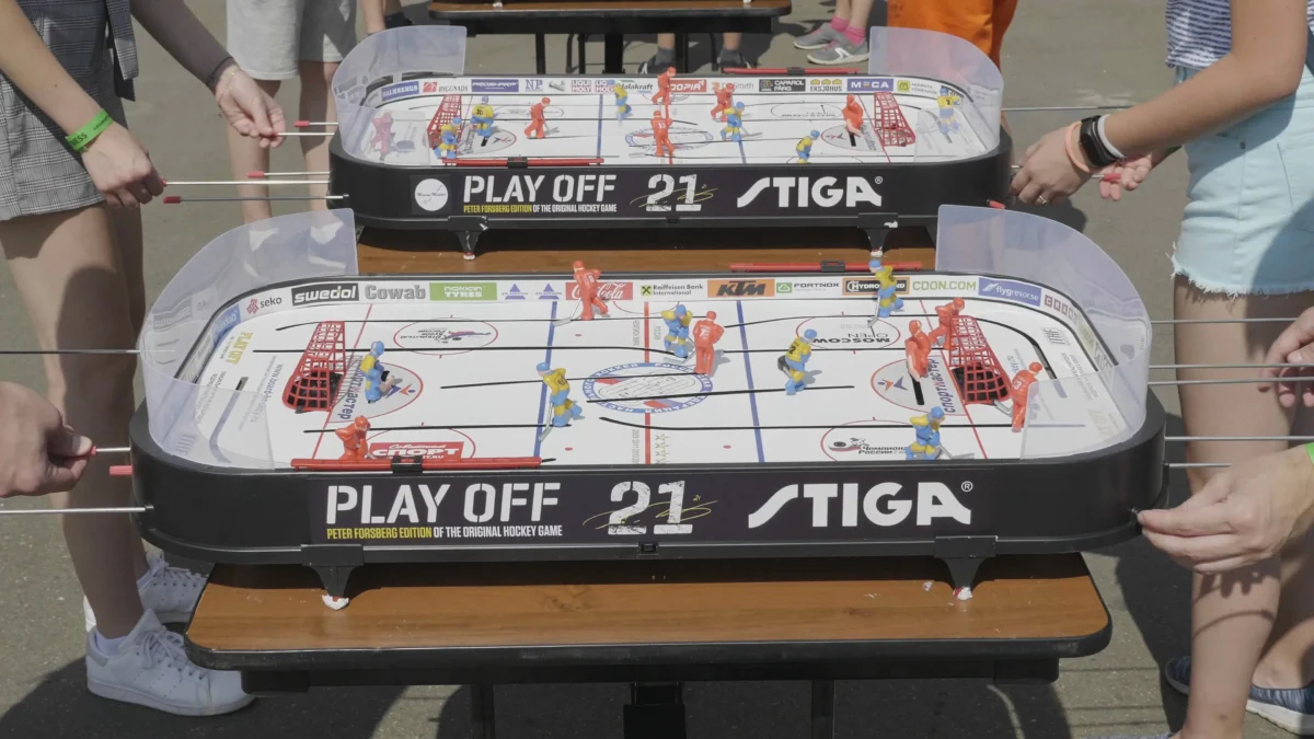 shutterstock_1662058048 1 | TVGURU.cz Tabletop hockey game with red and blue miniature players on a rink, surrounded by people holding rods and watching.