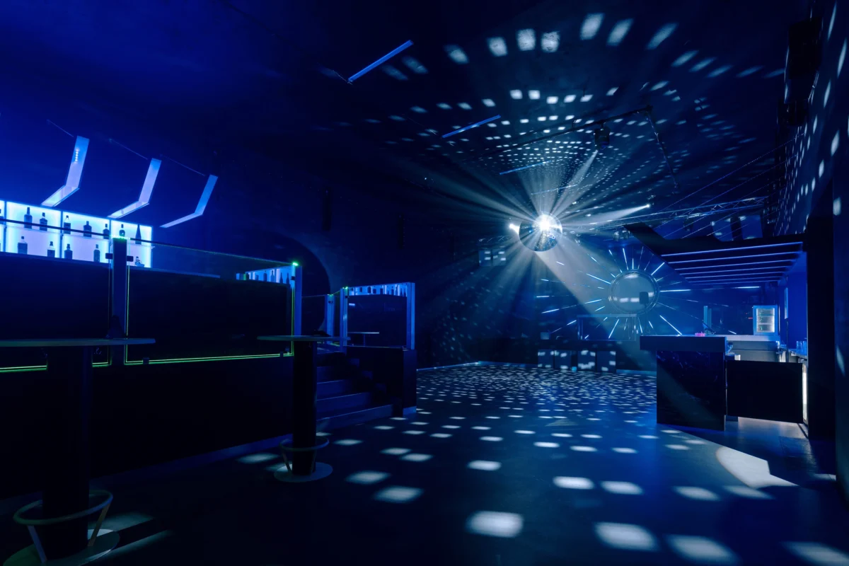 Blue-lit nightclub interior with a large disco ball projecting light dots across walls and floor, bar area on the left.
