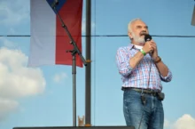 Older man with white beard singing into a handheld microphone on an outdoor stage, wearing a blue-and-white checkered shirt and jeans.