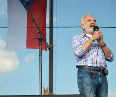 Older man with white beard singing into a handheld microphone on an outdoor stage, wearing a blue-and-white checkered shirt and jeans.