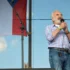 Older man with white beard singing into a handheld microphone on an outdoor stage, wearing a blue-and-white checkered shirt and jeans.