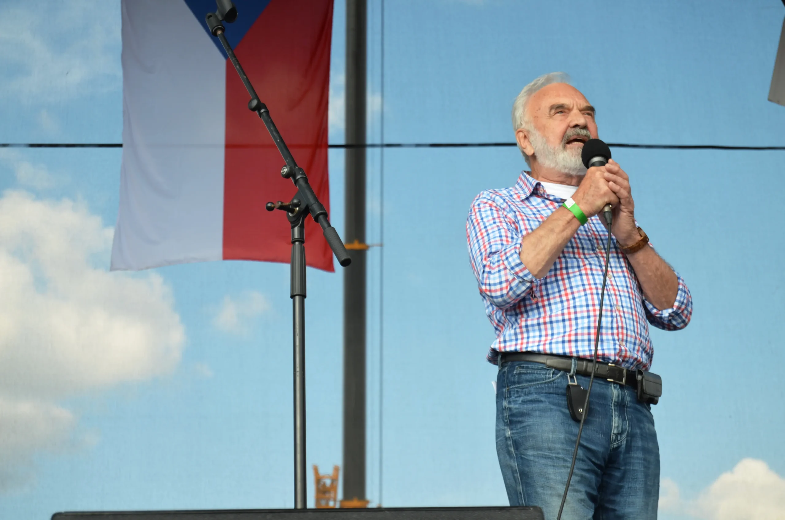 Older man with white beard singing into a handheld microphone on an outdoor stage, wearing a blue-and-white checkered shirt and jeans.